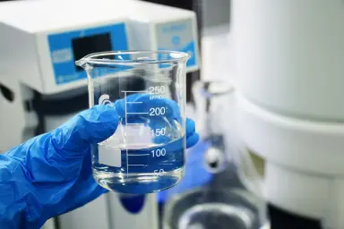Egy vegyianyag mérőedényt tartó laboráns kezének fotója